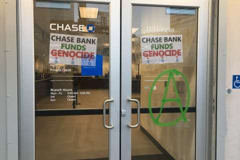 the front entrance to a chase bank with large plate glass doors. the doors have signs that read 'chase bank funds genocide'