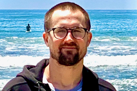 Chest-up photo of Casey - white, bespectacled, hair cut short, wearing a dark hoodie over a light T-shirt - at the beach.