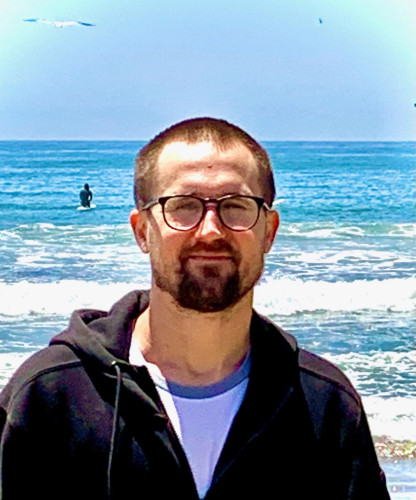 Chest-up photo of Casey - white, bespectacled, hair cut short, wearing a dark hoodie over a light T-shirt - at the beach.