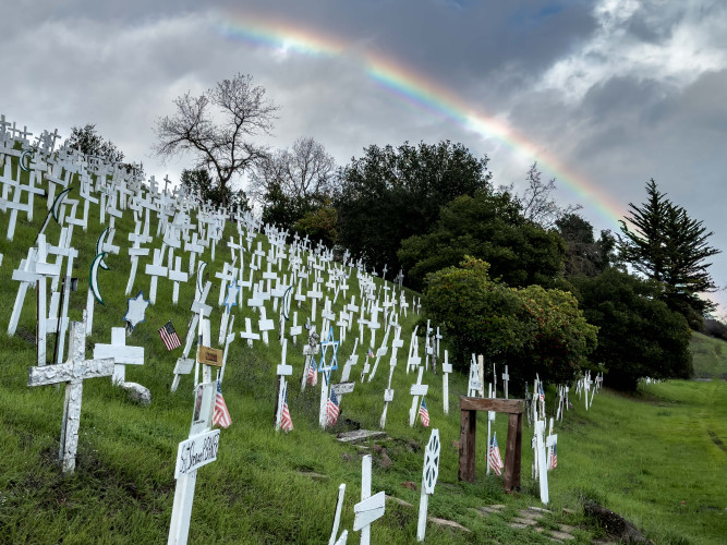 A rainbow arches above dozens of white crosses and symbols blanketing the hillside