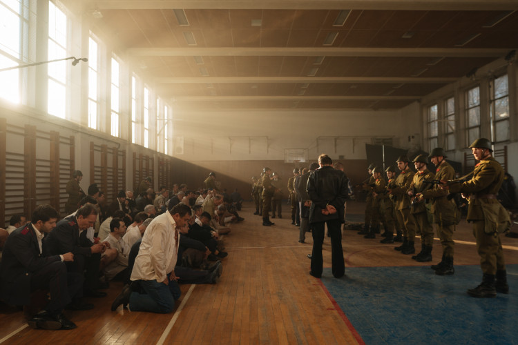 Multiple men in a gymnasium kneeling facing armed soldiers