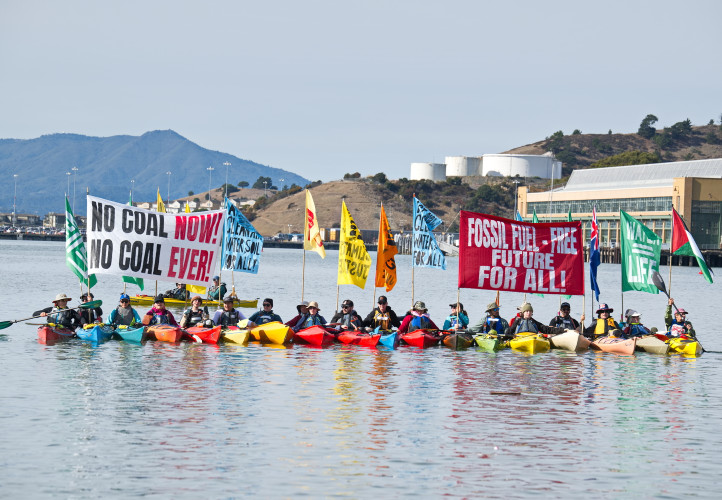 Kayaks sail with banners, flags and shouts that say “No Coal Coast to Coast,” “We’re Still Watching,” “Fossil Free Future for All,” “Newc...