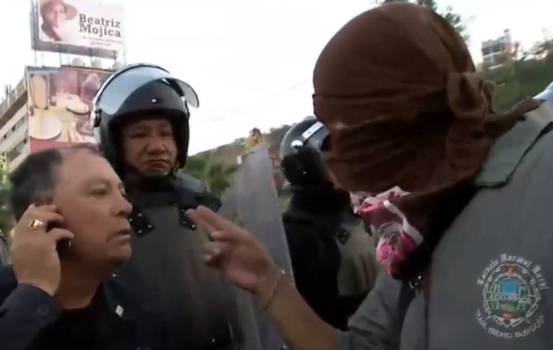 Person with face covered points two fingers at police officers.