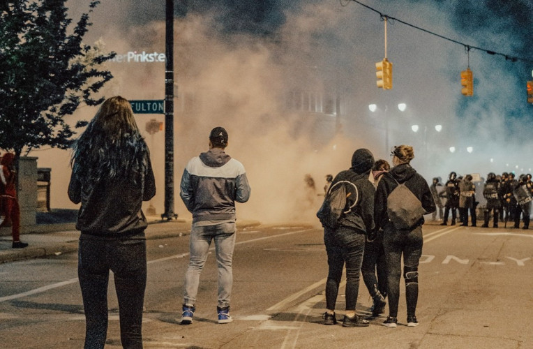 Six people are shown facing off against a line of cops standing in the middle of the street. Smoke fills the air.